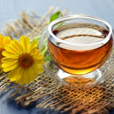A glass cup of honey with a yellow flower laying on the left. Both are on a piece of hessian and sunlight and reflections show through the glass