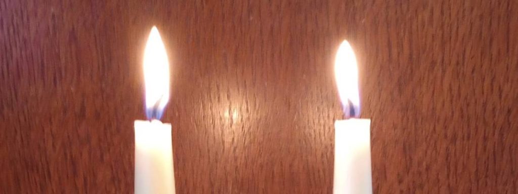 Two cream coloured beeswax candles alight with the flames contrasted against a brown wood background